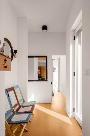 Un couloir avec deux chaises colorées, un plancher en bois, une porte blanche et un miroir accroché au mur.