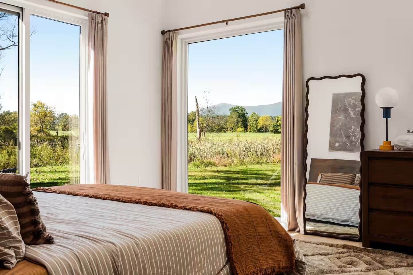 Ein Schlafzimmer mit einem großen Bett, einem Schminktisch, einem Spiegel und Blick auf eine grüne Landschaft durch ein Fenster.