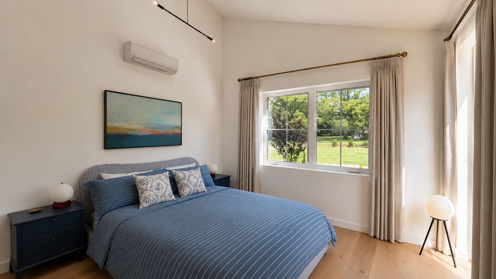 A bedroom with a bed, bedside tables, a nightstand, a painting, a window with curtains, and a view of greenery outside.
