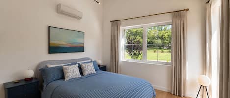 A bedroom with a bed, bedside tables, a nightstand, a painting, a window with curtains, and a view of greenery outside.