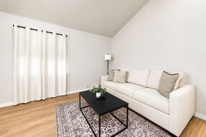 Een woonkamer met een witte bank, een zwarte salontafel met een plant, een tapijt met patroon en een witte muur met een zwarte gordijnroede.