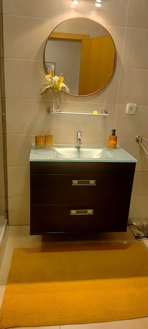 Ein Badezimmer mit einem dunklen Waschtisch, einem runden Spiegel, einem Glaskonstruktion-Waschbecken und einer gelben Badematte.