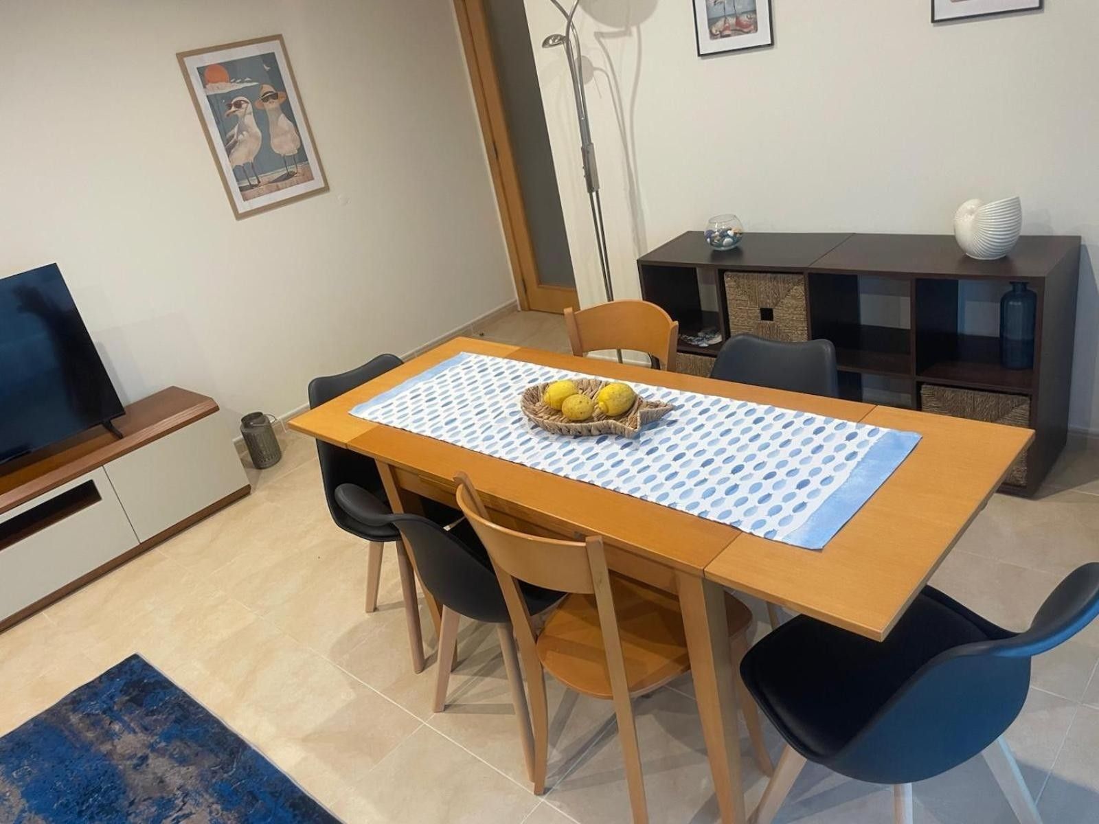 A dining area with a table, chairs, a fruit basket, and a television on a stand.