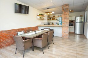 Een eetgedeelte met een tafel met glazen blad en rieten stoelen, een keuken met een waterdispenser en een koelkast, en een accentmuur van baksteen.