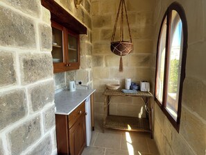 A stone-walled kitchen with a wooden cabinet, a hanging basket, and a window with a view. - Nostalgia suite at Dar Tal Kaptan, Ghasri, Gozo. Victorian themed room (Ghasri)