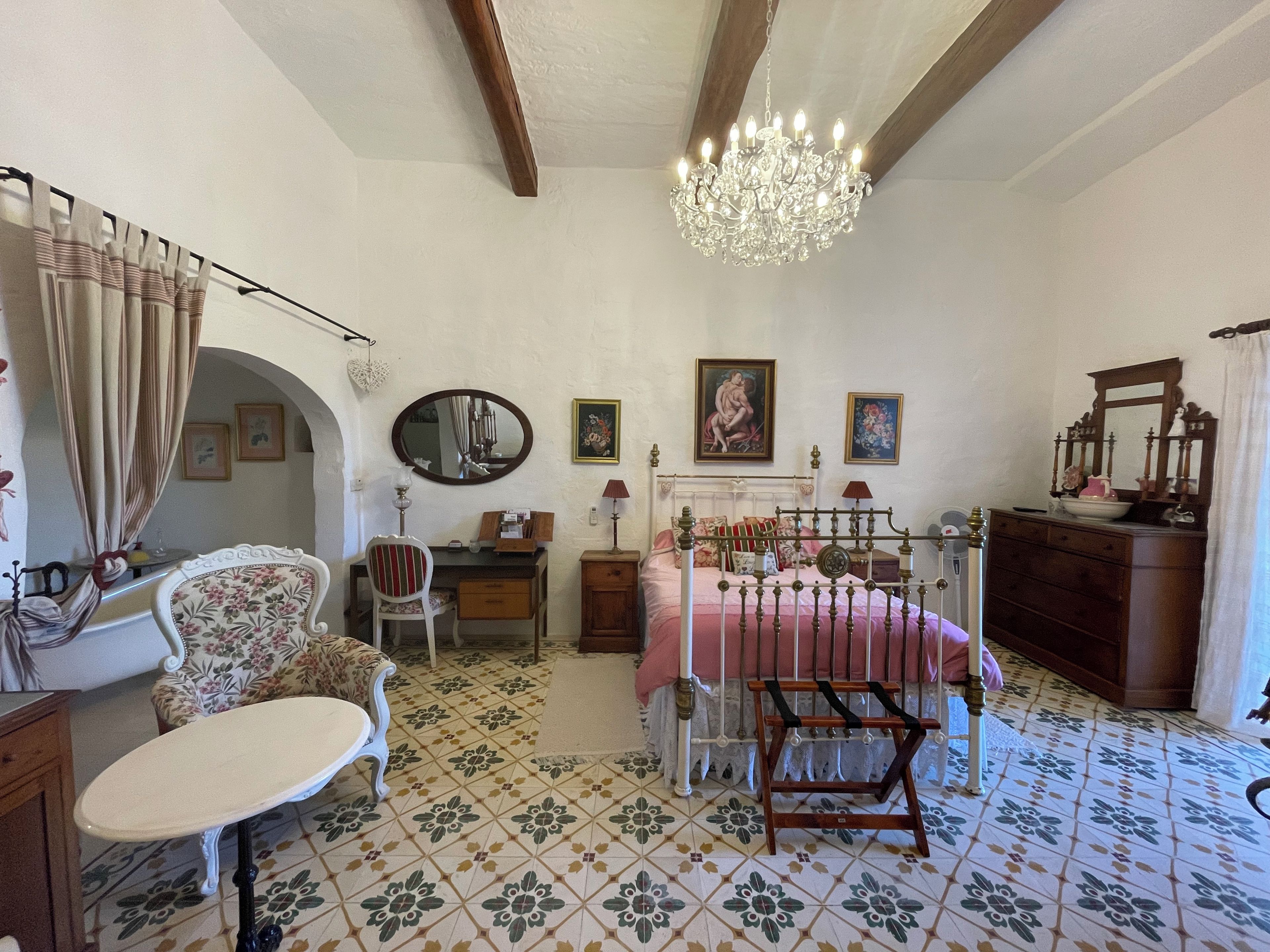 A bedroom with a bed, a chair, a desk, a dresser, a mirror, and a chandelier.