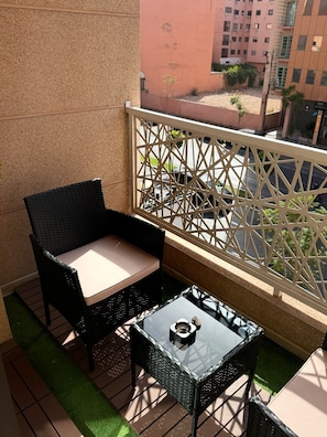 Ein Balkon mit Korbstuhl, kleinem Tisch und einem dekorativen Metallzaun, im Hintergrund ein Blick auf ein Gebäude.
