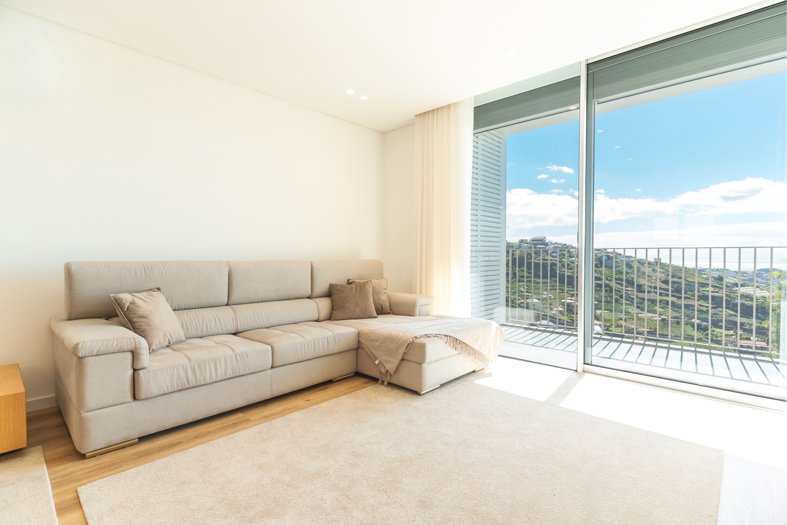 Ein modernes Wohnzimmer mit einem beigefarbenen Ecksofa, Holzboden und einer großen Schiebeglastür, die auf einen Balkon mit Blick auf die Stadt führt.