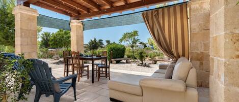 Eine Außenpatio mit einer Holz-Pergola, Steinsäulen, einem beigen Sofa und einem Esstisch mit Stühlen.