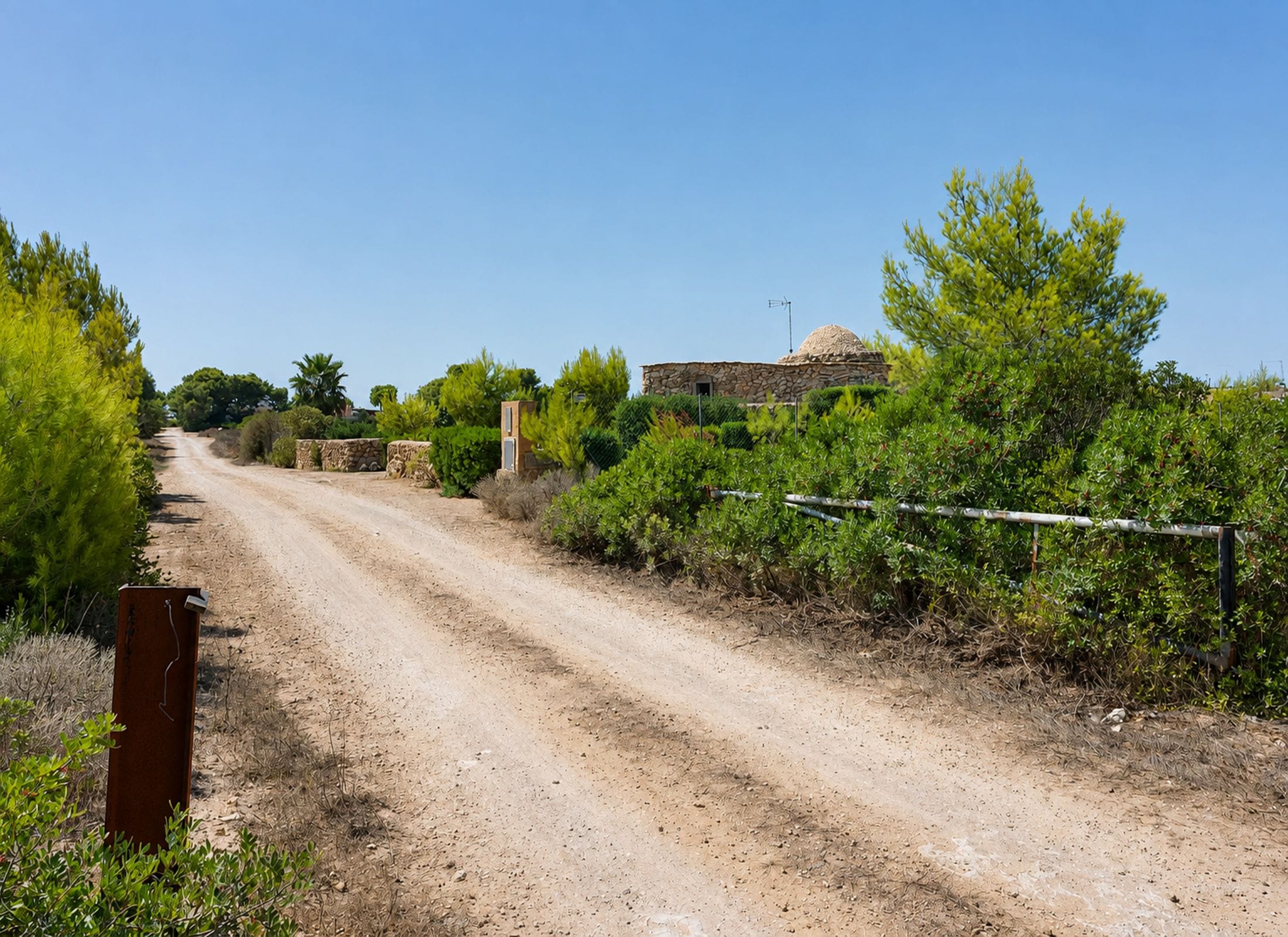 A rural dirt road flanked by greenery and a stone building.