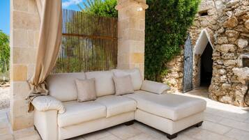 Un divano angolare beige con cuscini, posizionato sotto una pergola in legno con pannelli in bambù, su uno sfondo di muro in pietra e vegetazione.
