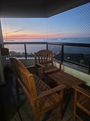 A balcony with wooden furniture, a view of the sea, and a sunset.