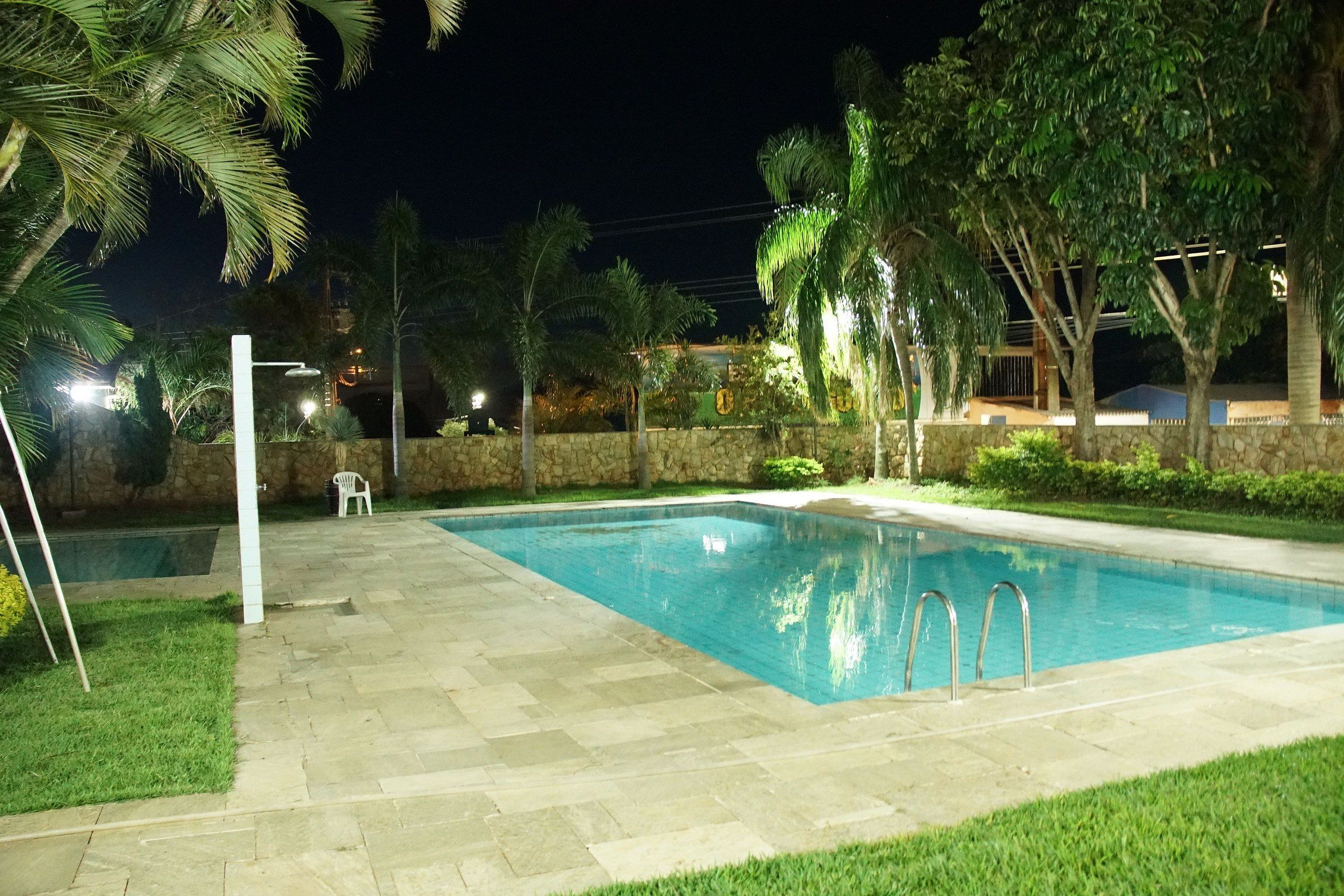 A swimming pool with a stone border, surrounded by grass and palm trees, with a stone wall and a chair in the background.