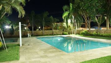 A swimming pool with a stone border, surrounded by grass and palm trees, with a stone wall and a chair in the background.