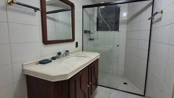 Uma casa de banho com soalho em azulejos brancos, toucador de madeira com espelho, cabine de duche e iluminação no teto.