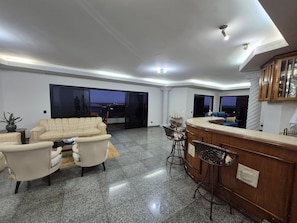 A spacious living area with a sofa, chairs, a bar area, and a view through large windows.