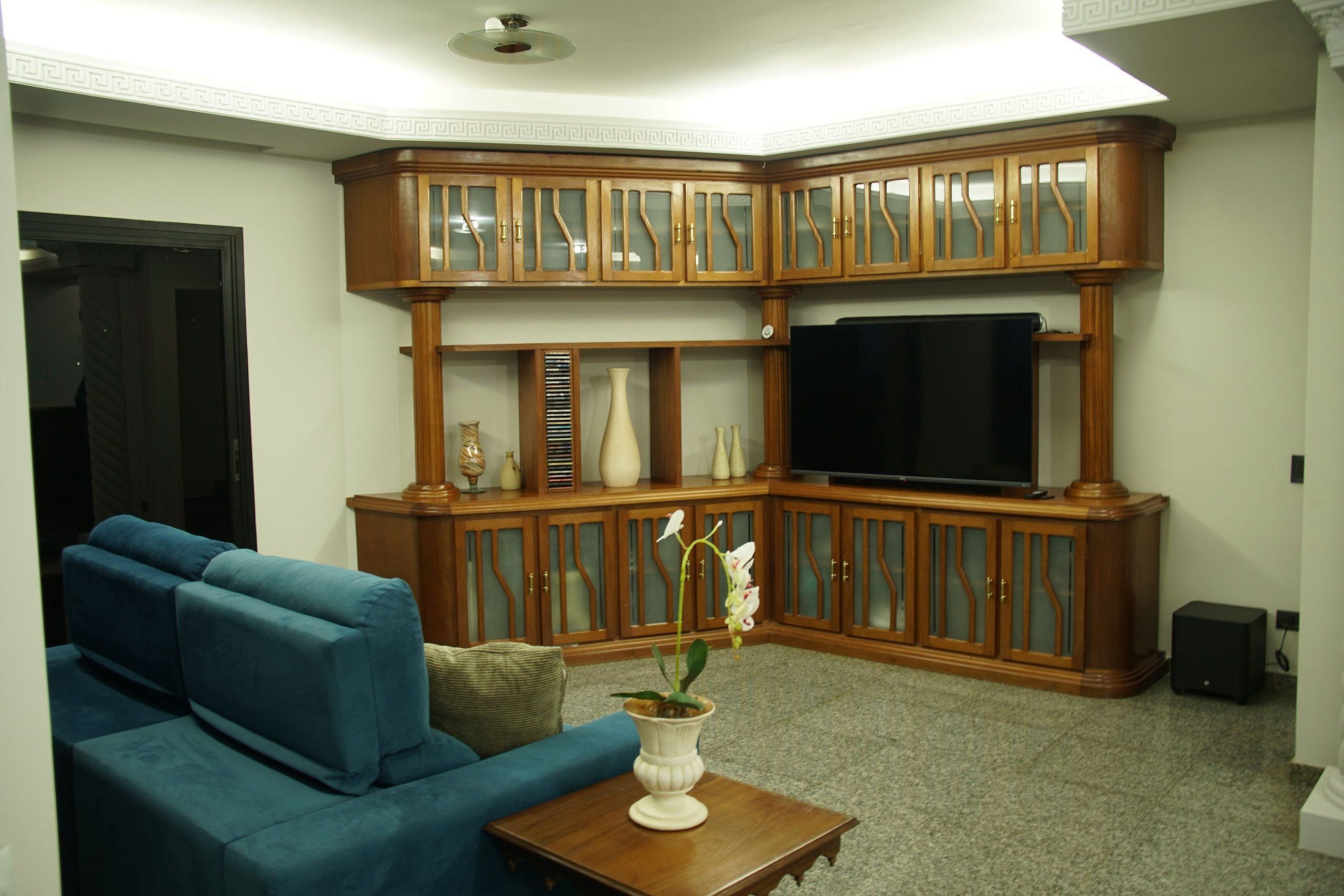 Una sala de estar con un sofá modular, un televisor, armarios de madera y un jarrón con flores.