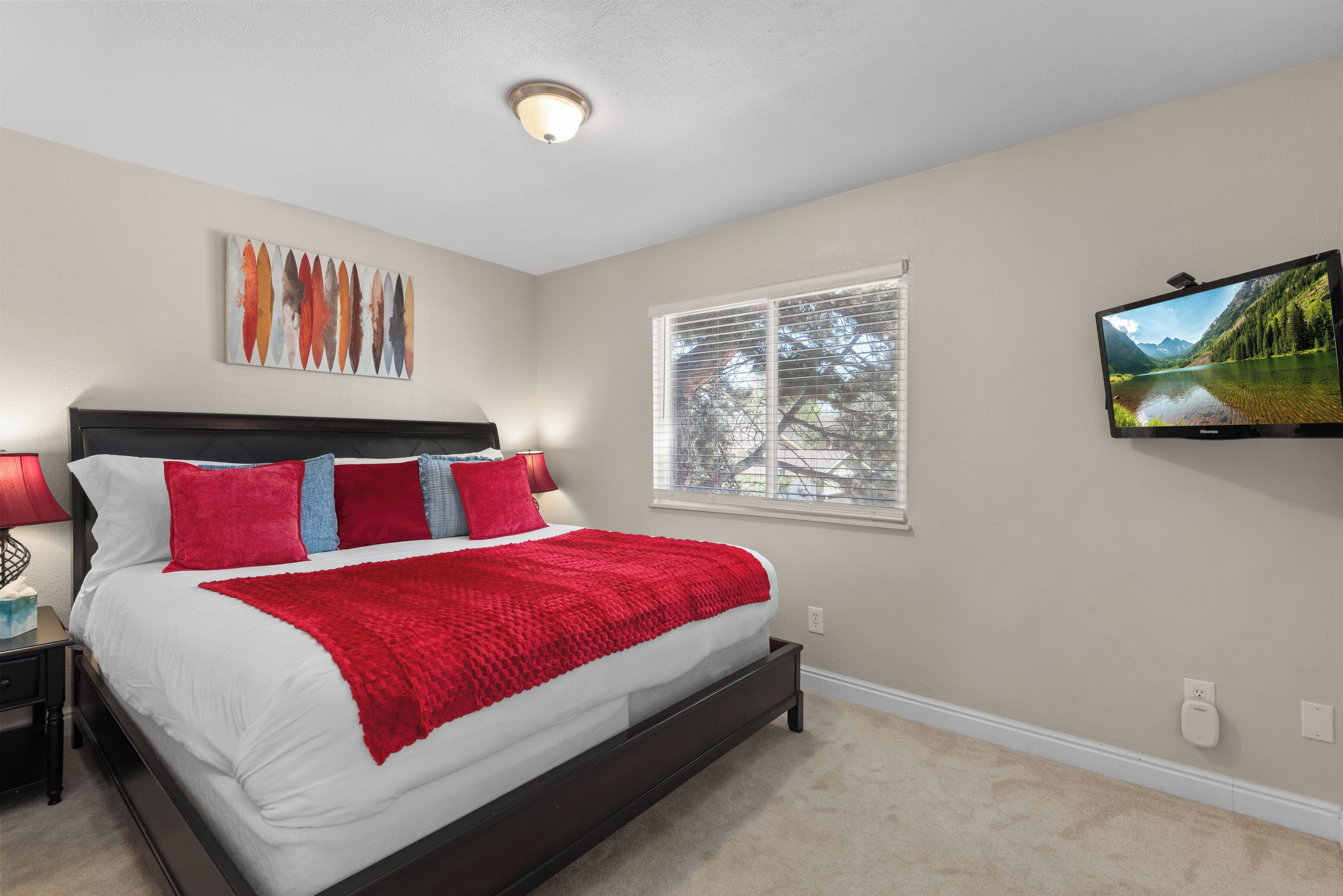 A bedroom with a bed, a nightstand, a lamp, a painting, a window with blinds, and a television on the wall.