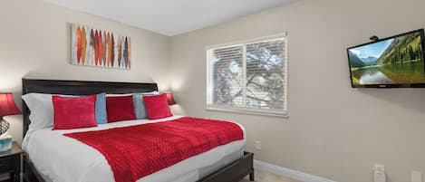 Un dormitorio con cama, mesita de noche, lámpara, cuadro, ventana con persianas y televisor en la pared.