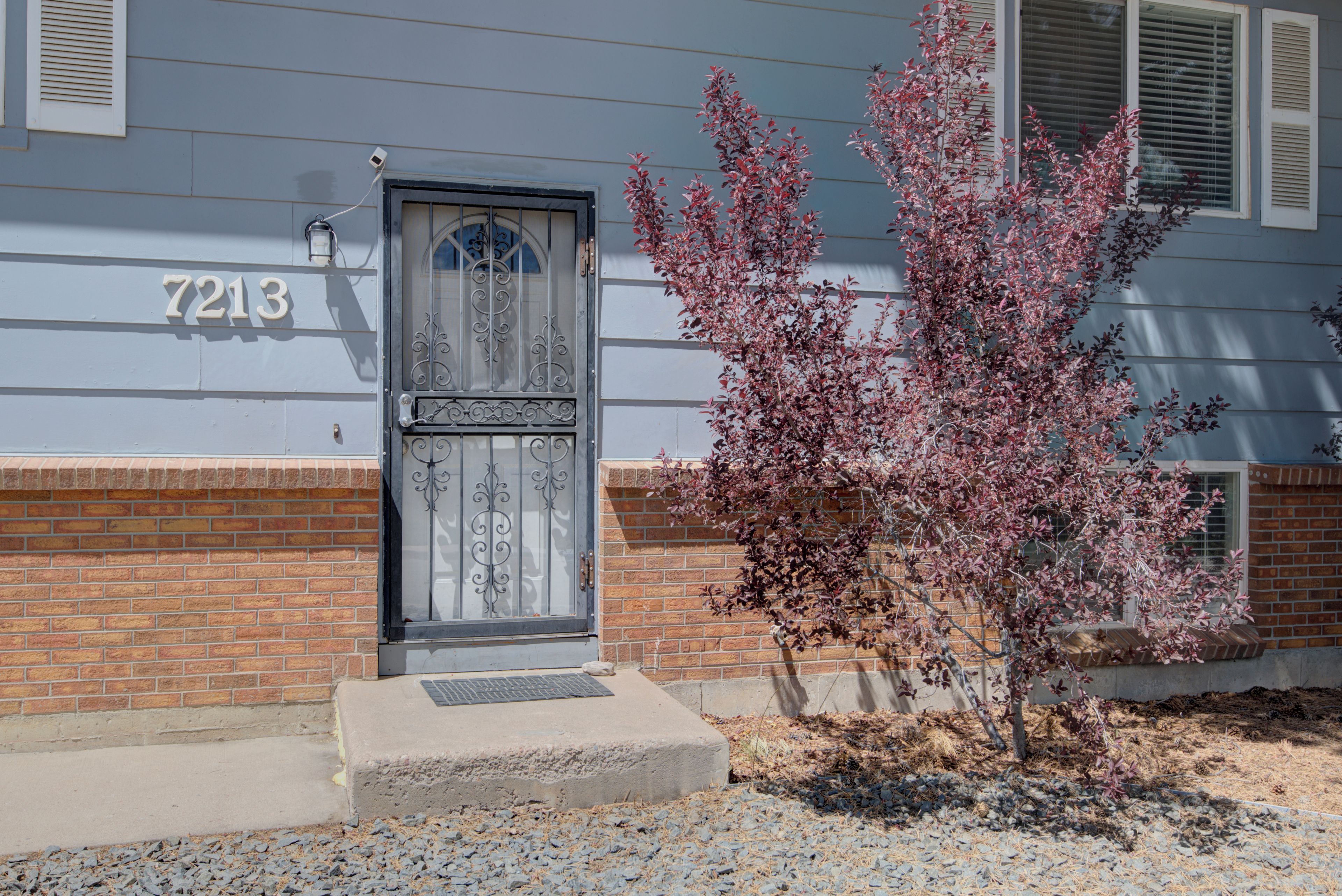 A house with a number 7213, a black door with decorative ironwork, and a small tree with pink leaves.
