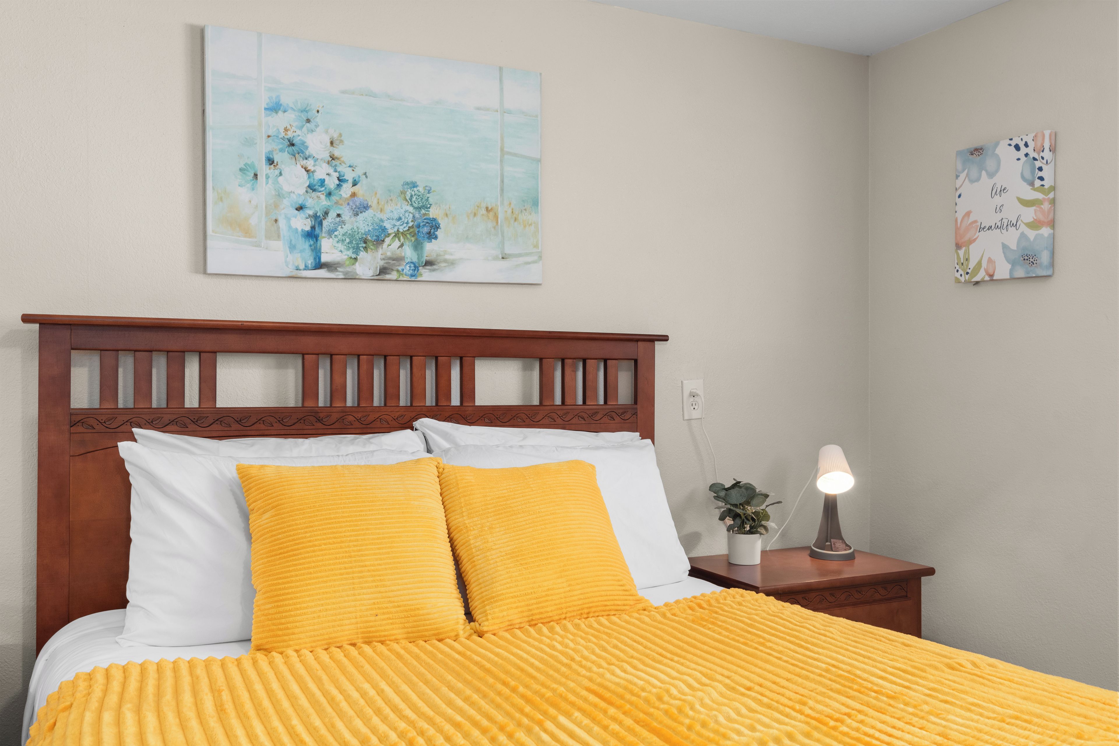 Un dormitorio con cabecera de madera, cama con ropa de cama amarilla, una mesita de noche con lámpara y dos obras de arte en la pared.