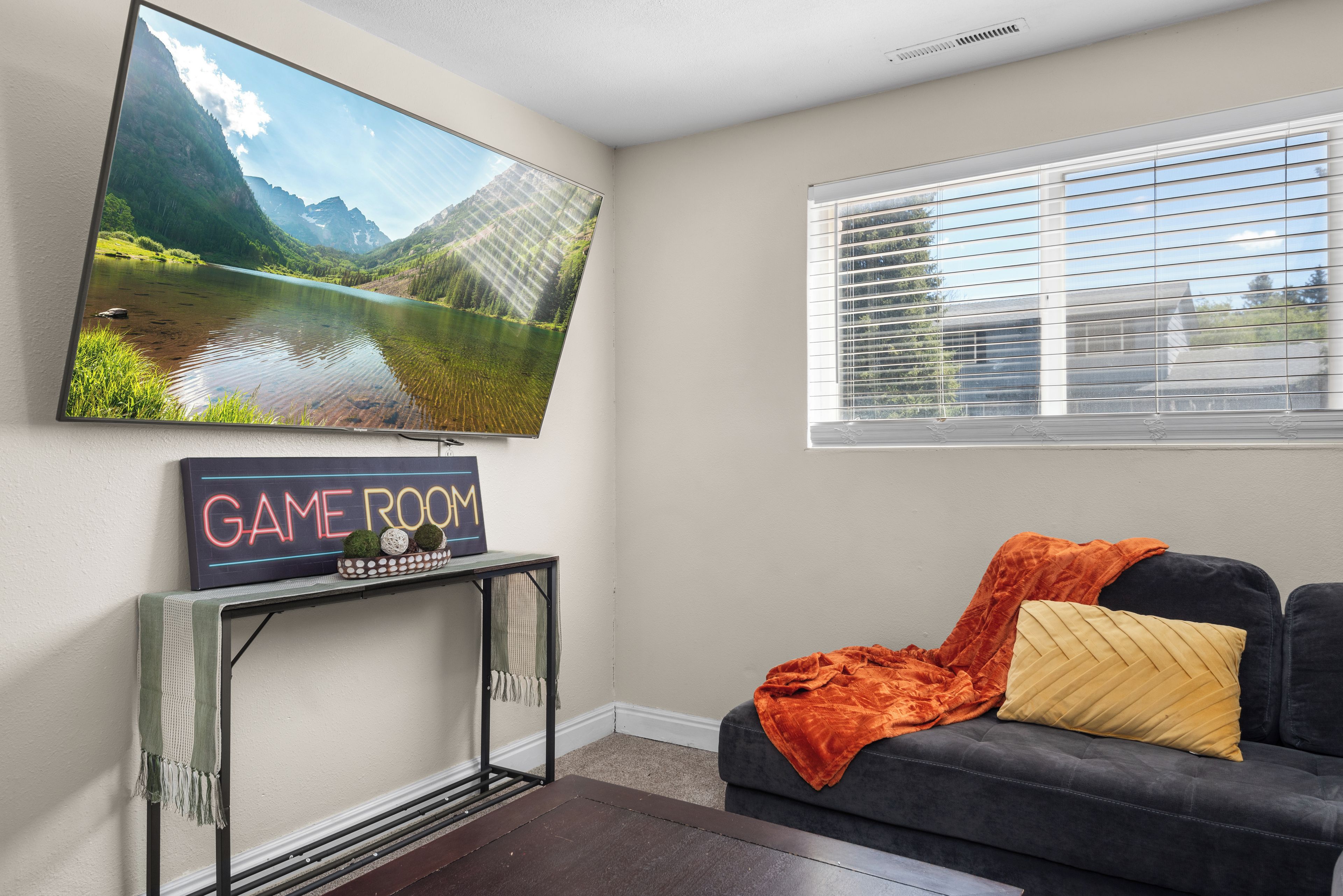 A living room with a flat-screen TV mounted on the wall, a Game Room sign, a dark sofa with cushions, and a window with blinds.