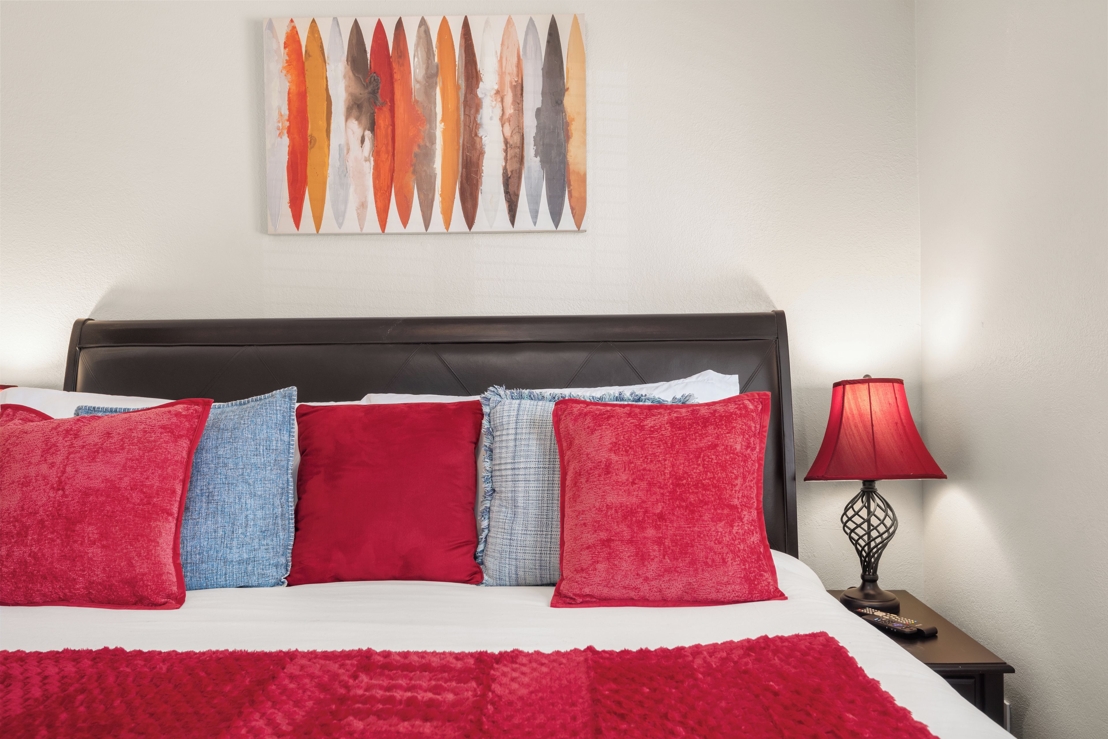 Una cama bien hecha con almohadas rojas y azules, una manta roja y un cuadro abstracto colgado en la pared.
