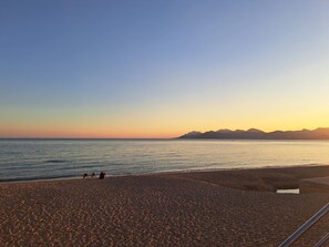 Ein Strand mit blauem Himmel und ruhiger See, wenige Personen im Sand und entfernte Berge.