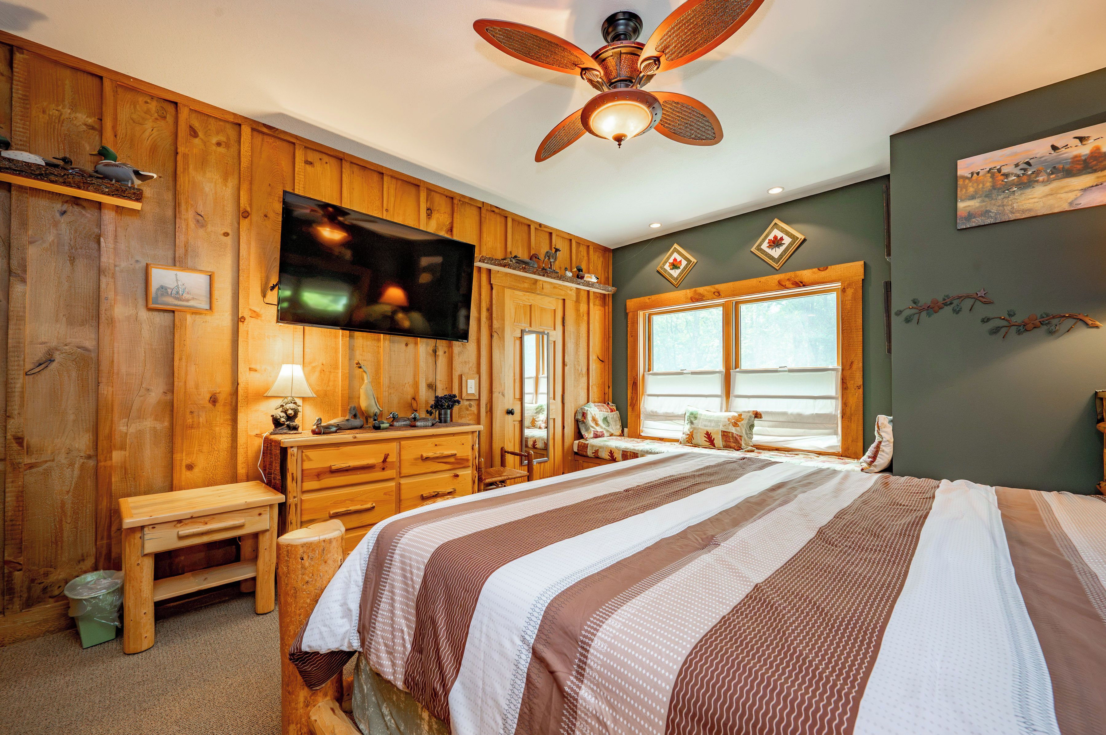 A room with a bed, wooden walls, a ceiling fan, a television, and a window with a view.