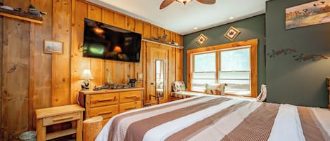 A room with a bed, wooden walls, a ceiling fan, a television, and a window with a view.