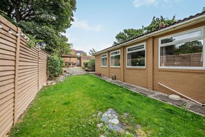Un jardin à l’arrière d’une maison, avec une pelouse, une clôture en bois et une maison dotée de grandes fenêtres.