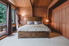 A bedroom with a wooden headboard, a bed with patterned bedding, two bedside lamps, and a view of the outdoors through a large window.