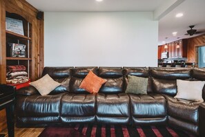A leather sofa with multiple cushions, a wooden bookshelf with magazines, and a kitchen area in the background.