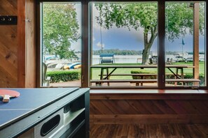 A room with a ping pong table, large windows with a view of a lake, picnic tables, and a tree.