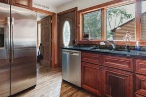 A kitchen with wooden cabinets, a stainless steel refrigerator, a dishwasher, and a window with a view of trees. - Long Lake Extended-Stay Furnished 30-Day+ Home (Ingleside)