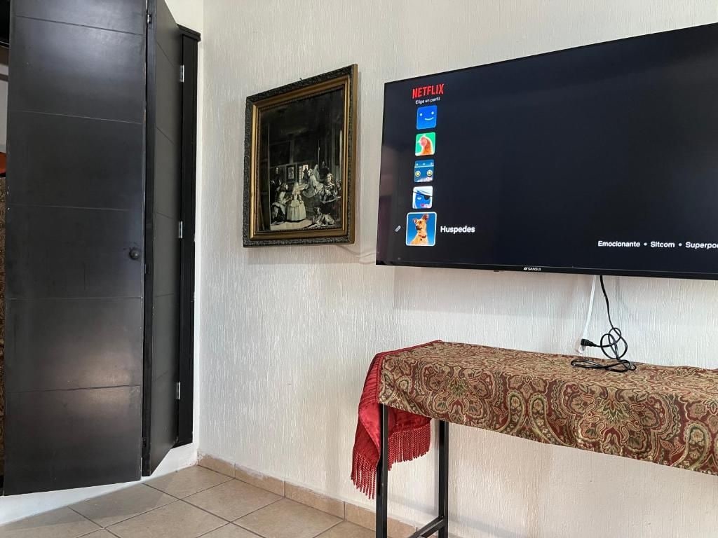 Una stanza con una televisione fissata al muro che mostra l'interfaccia di Netflix, un quadro incorniciato sopra e un tavolo con una tovaglia fantasia.