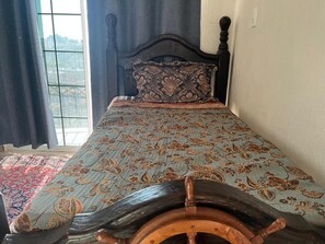 Una cama con una colcha estampada y un cojín decorativo, un cabecero de madera y vistas a través de una ventana.