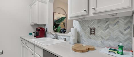 A modern kitchen with white cabinetry, a granite countertop, a coffee maker, and a herringbone tile backsplash.
