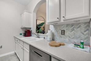 A modern kitchen with white cabinetry, a granite countertop, a coffee maker, and a herringbone tile backsplash. - Balcony Bliss At Windsor Hills (Kissimmee)