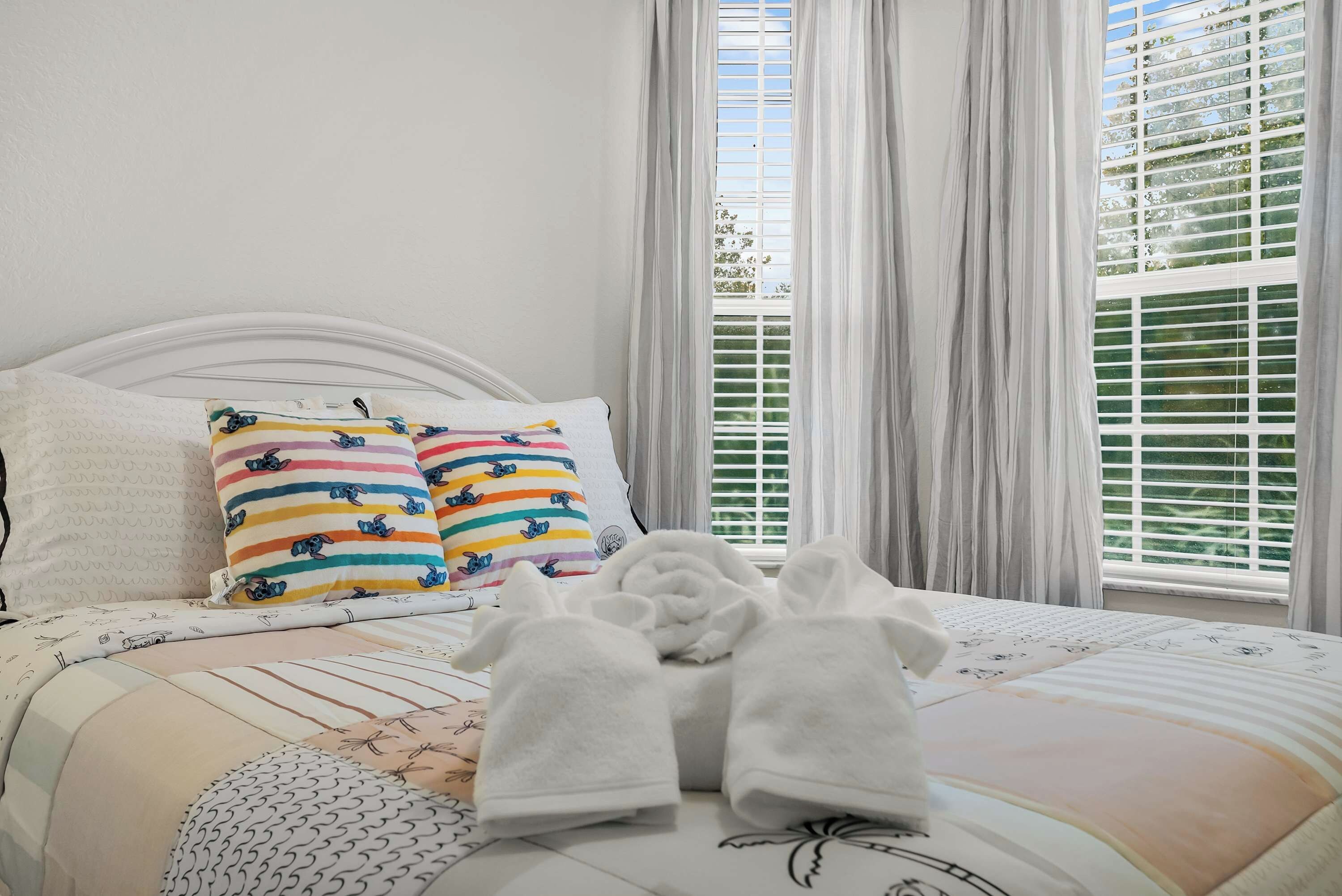 Un lit bien fait, avec du linge de lit blanc et deux coussins colorés, une fenêtre avec des stores et une vue sur la verdure à l’extérieur.