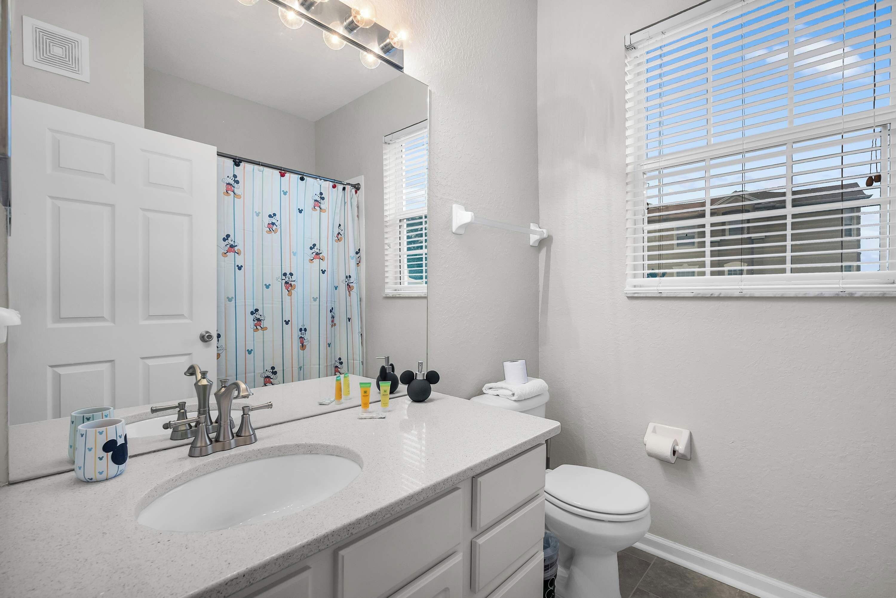 Une salle de bain avec un lavabo, un meuble-lavabo et des toilettes, tous de couleur blanche. On y trouve un rideau de douche à l’effigie de Mickey Mouse, un miroir avec éclairage intégré et une fenêtre avec des stores.