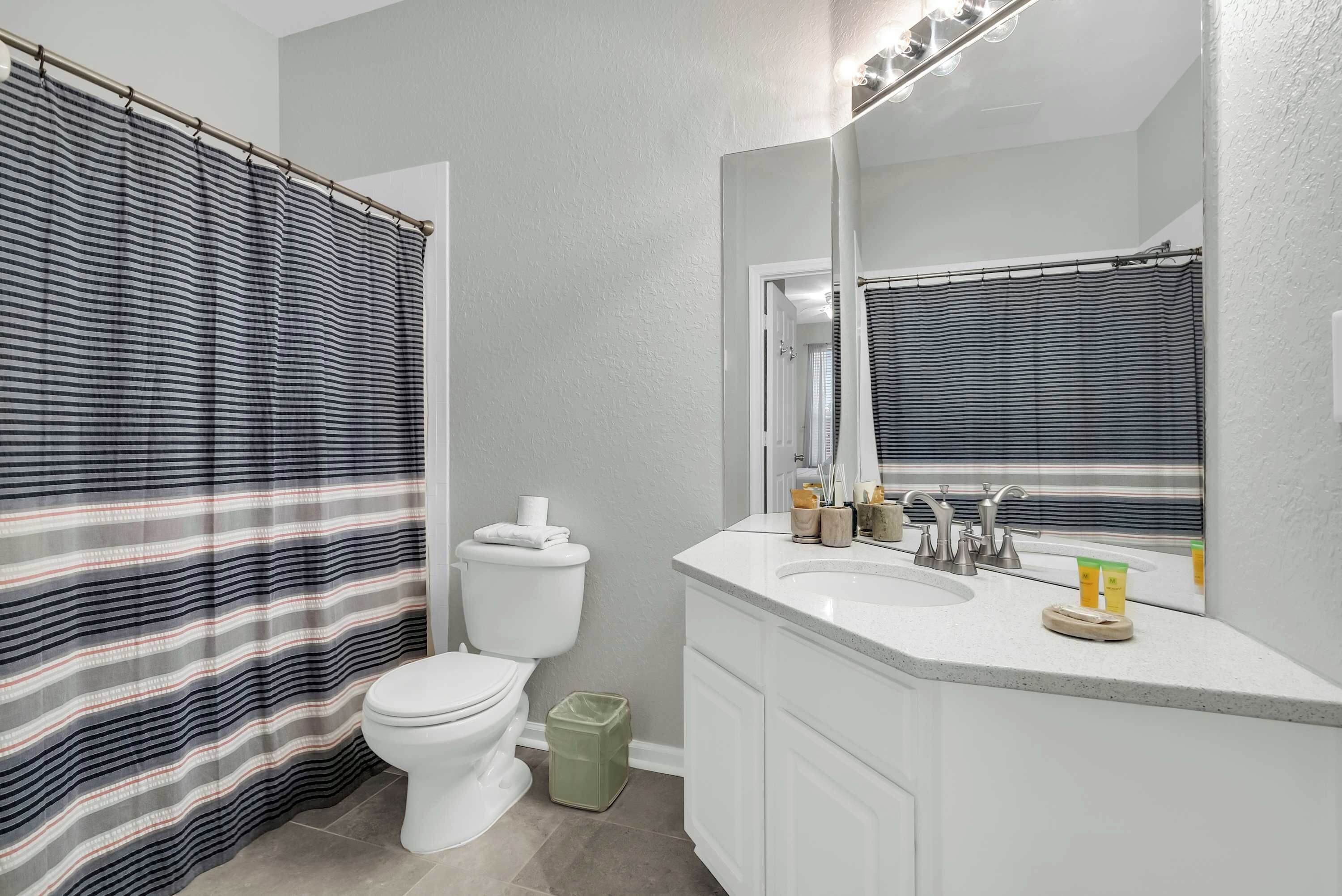Une salle de bain avec un rideau de douche à rayures, des toilettes, un lavabo avec un miroir au-dessus et une poubelle.