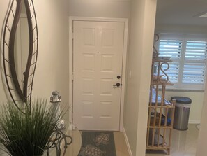 A hallway with a white door, a mirror, a potted plant, a small table with a decorative item, and a window with blinds. - Cozy 2-bedroom and 2 bath condo with AC in terrific Fort Myers (Fort Myers)
