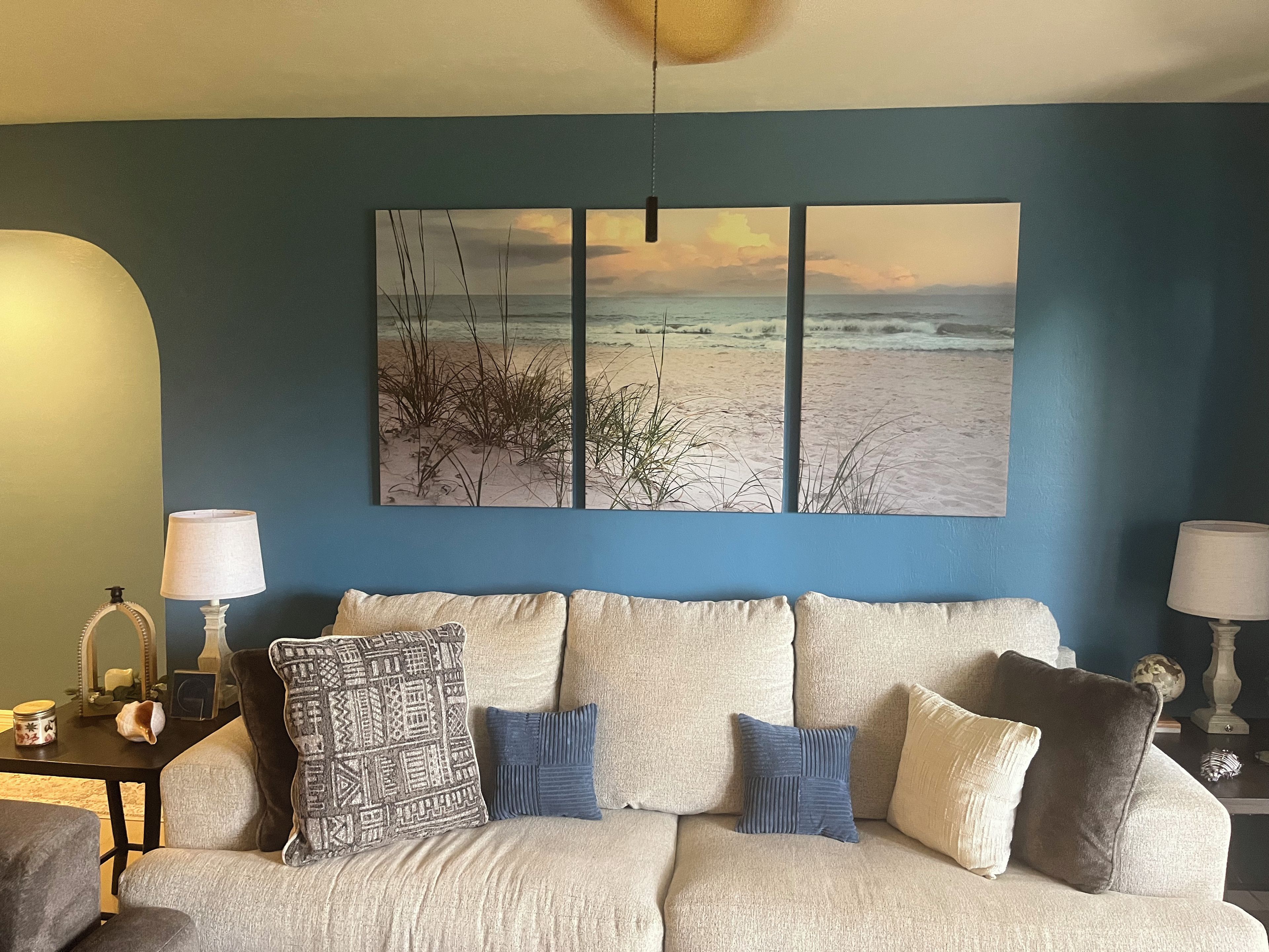 A living room with a large beach-themed artwork on the wall, a beige sofa with multiple cushions, and two side tables with lamps.