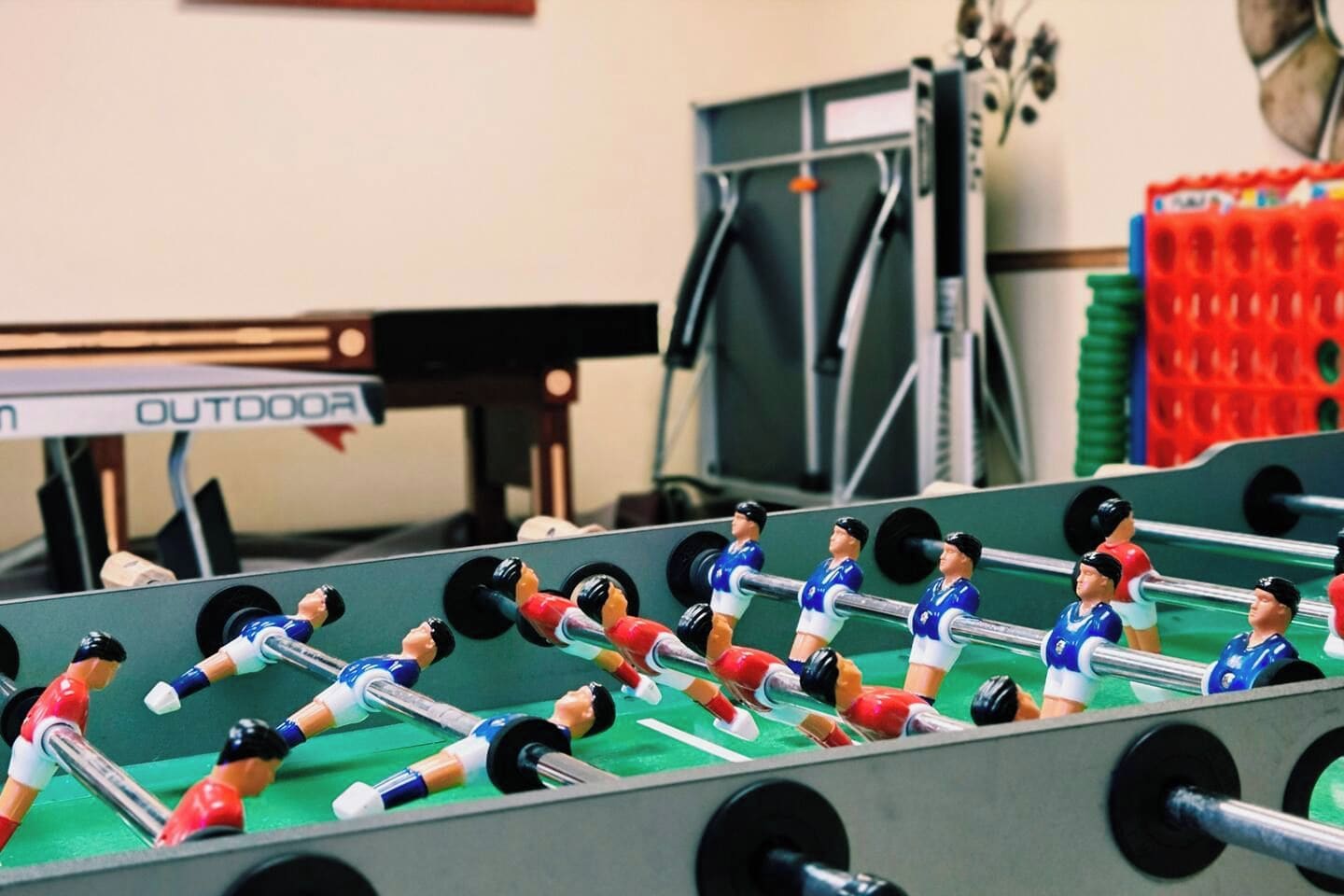 Een tafelvoetbaltafel met meerdere spelers en een pingpongtafel op de achtergrond.