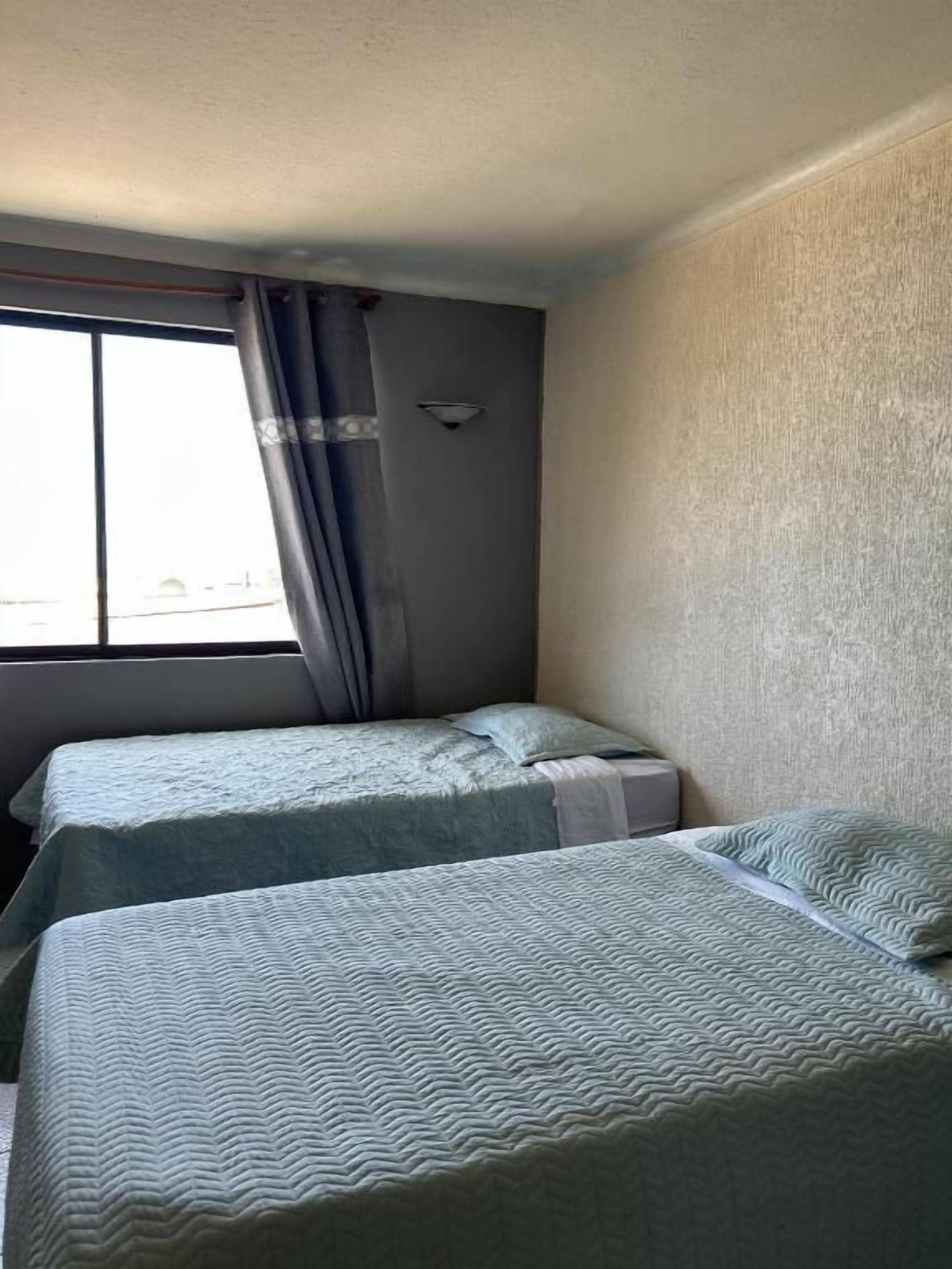 Una habitación con dos camas, un ventanal con cortinas y una lámpara en la pared.
