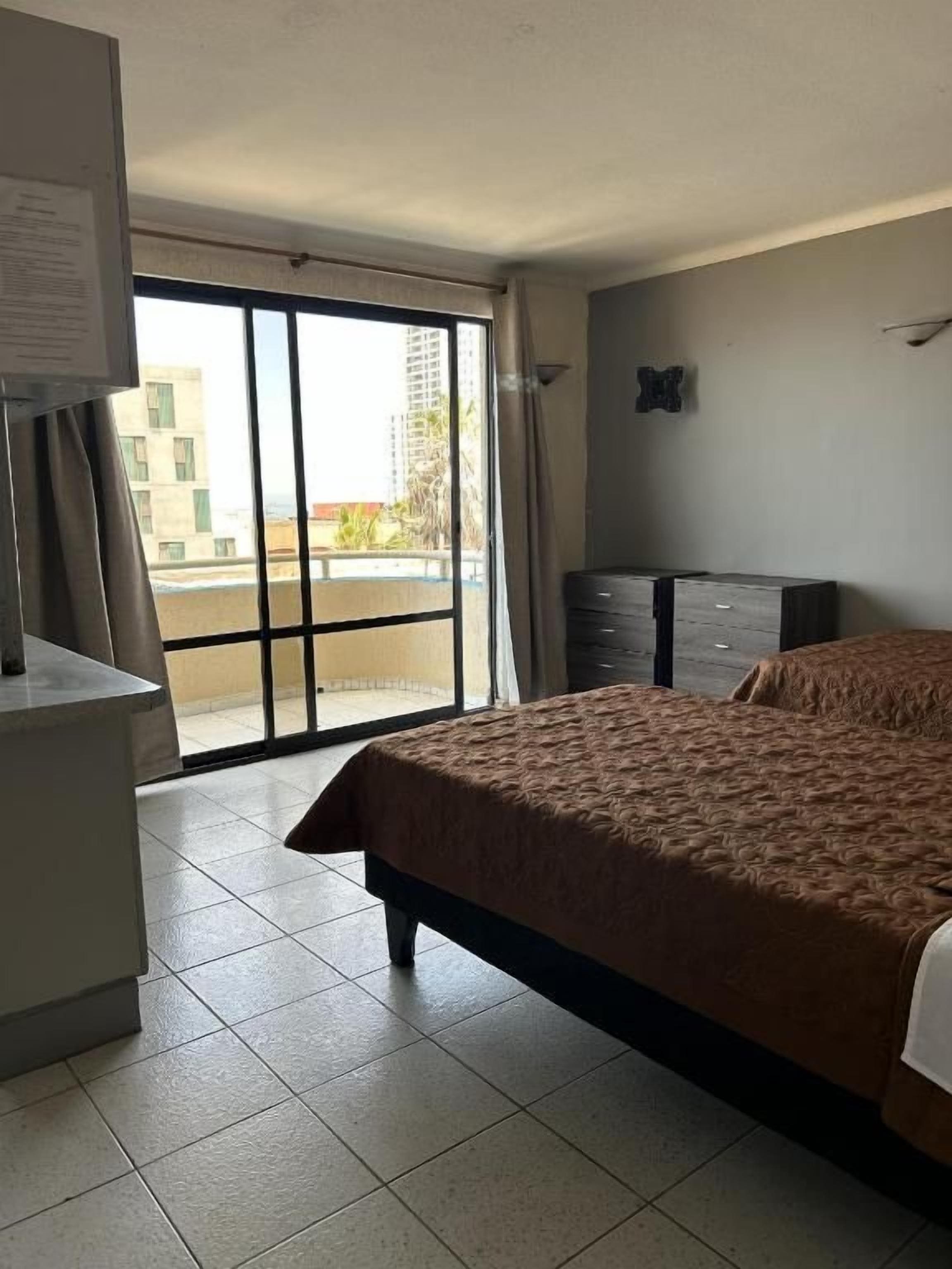 A hotel room with a bed, a dresser, a desk, and a sliding glass door leading to a balcony.