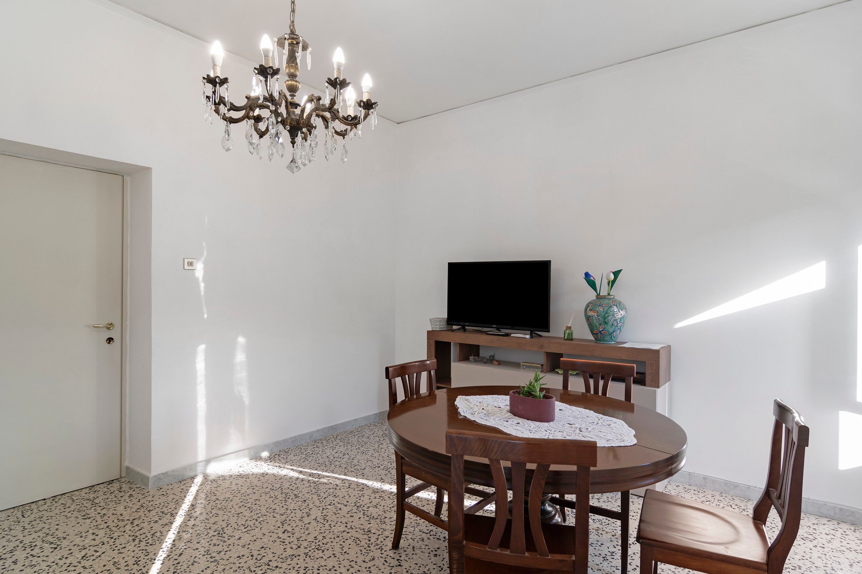 A dining area with a round table, chairs, a chandelier, a flat-screen TV, and a decorative vase.