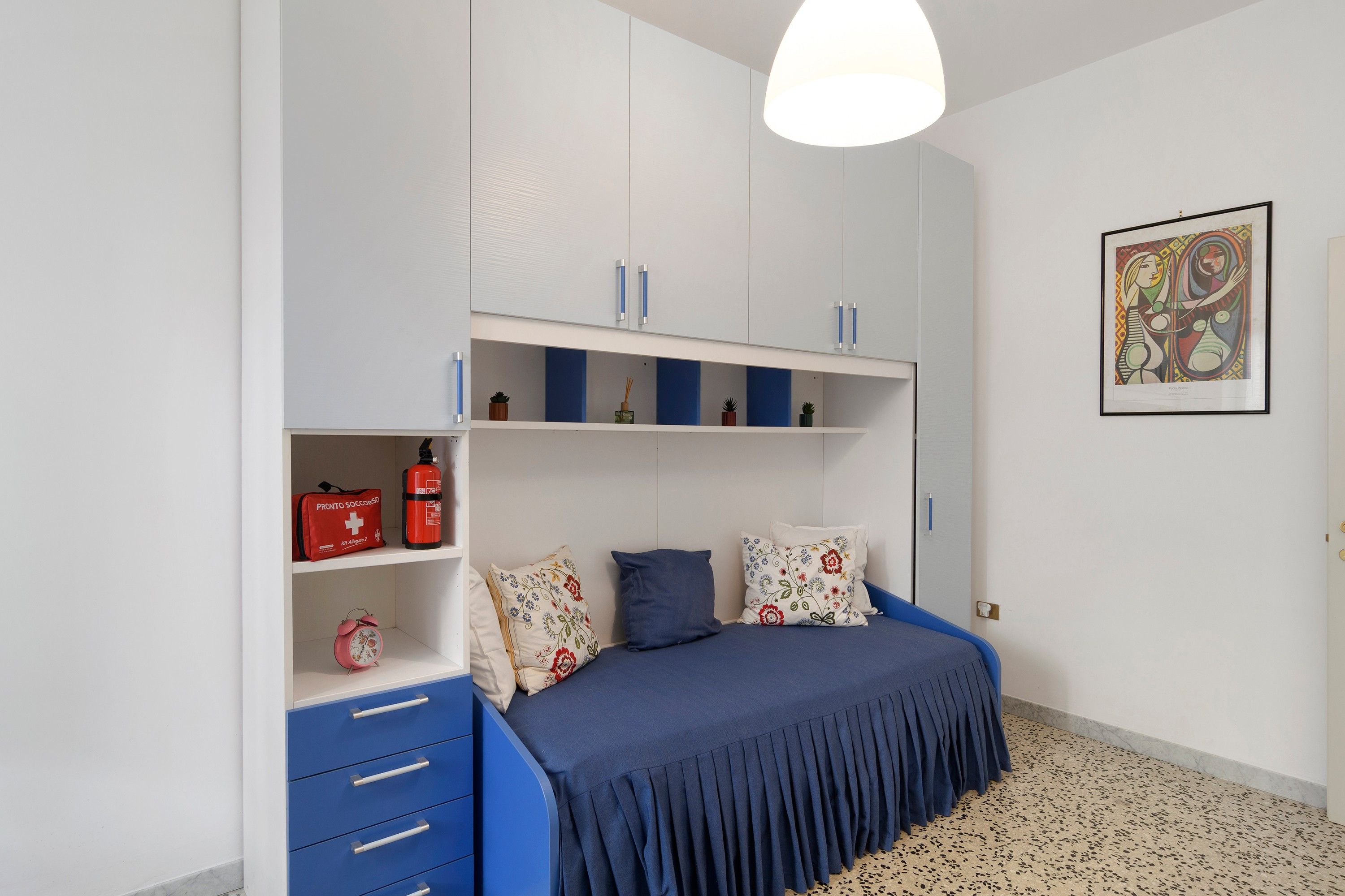 Una camera da letto con un letto, cassetti, una mensola con oggetti e un quadro appeso al muro.