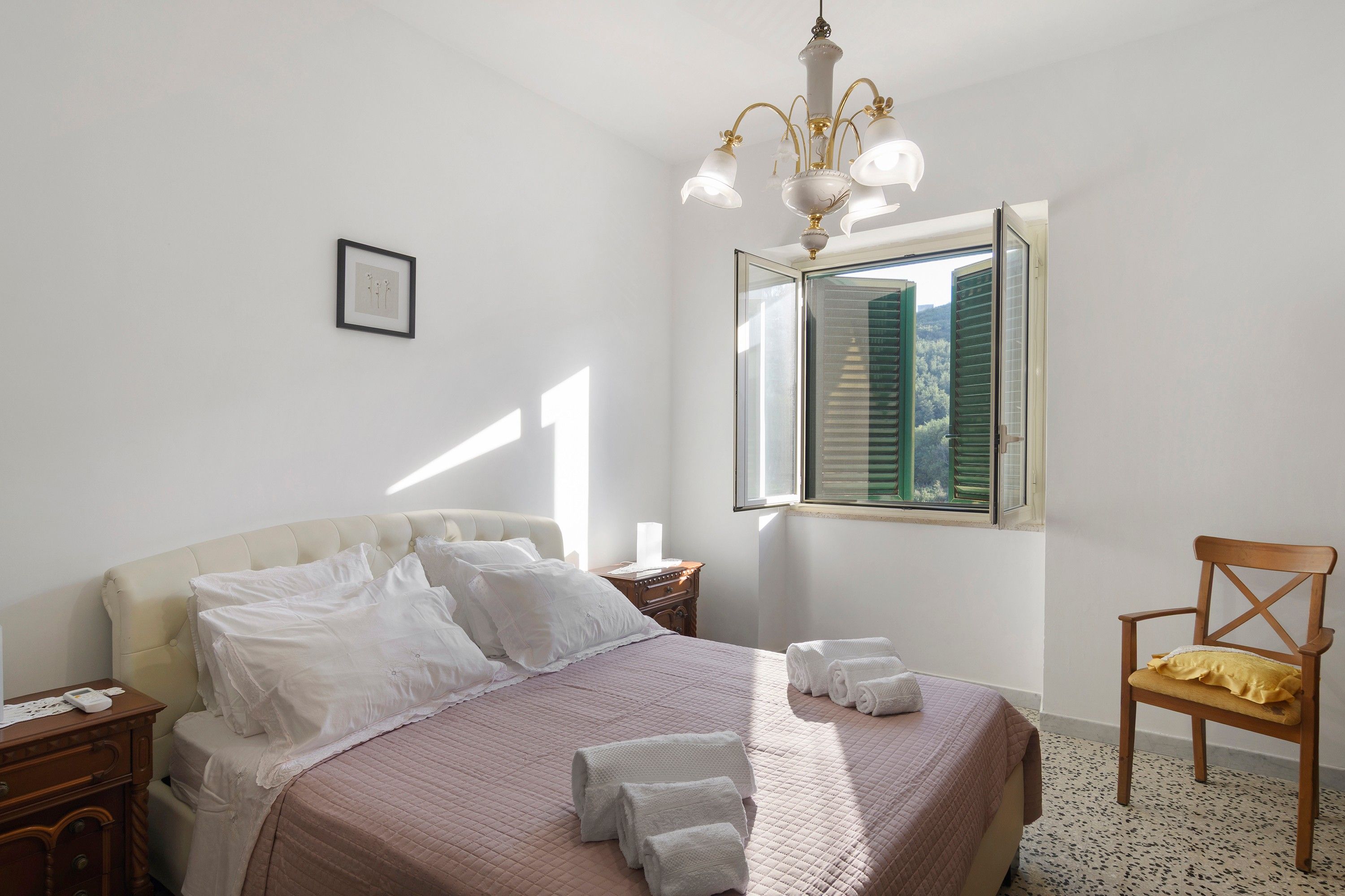 A bedroom with a bed, a nightstand, a chair, a window with shutters, and a chandelier.